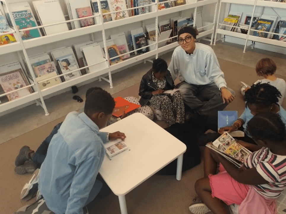 Iara MONTEIRO BRADAO, bénévole à l'association Axes pluriels, assise avec des élèves de primaires à la bibliothèque municipale en train lire des livres