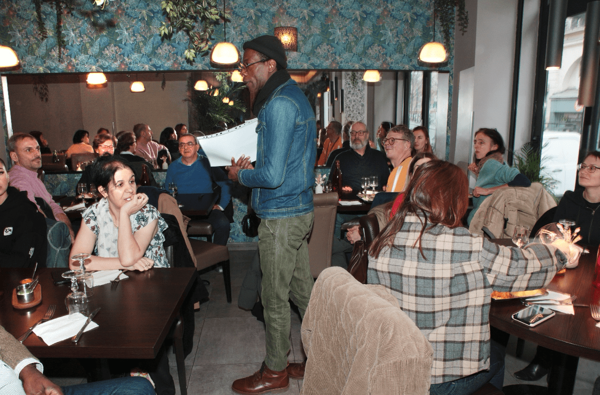 Vue larges des personnes assises à table présentes avec Charles Kingue débout au centre lors du repas de fin d'année 2025 de l'association Axes Pluriels